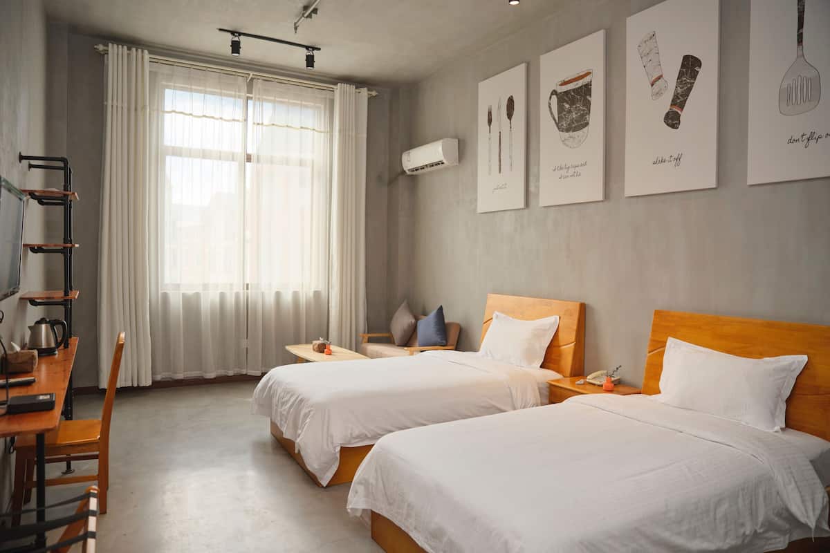 Habitación de hotel con dos camas, un escritorio con silla, una mesita y una ventana con cortinas. Se observan obras de arte colgadas en la pared y un equipo de aire acondicionado.