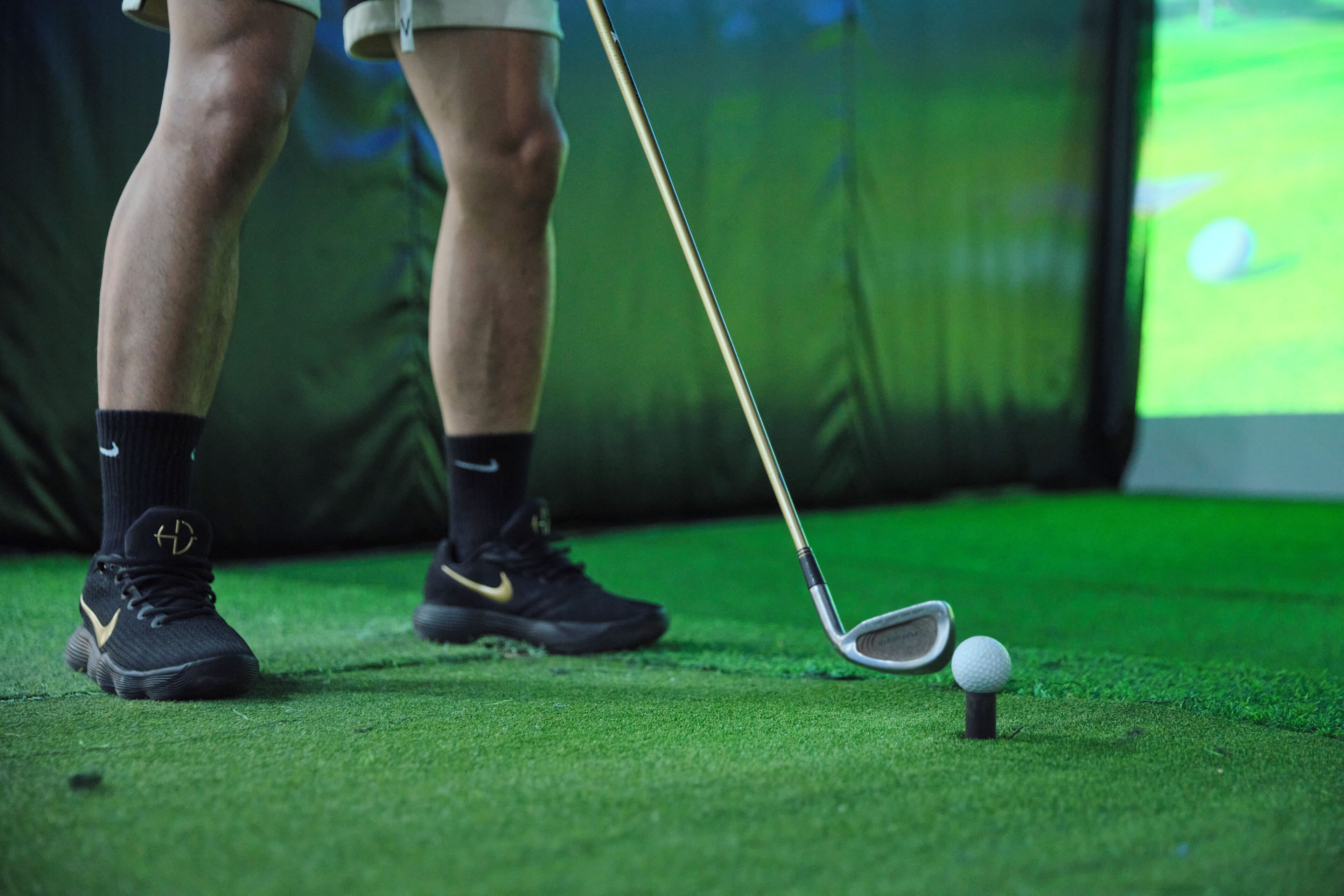 Een detailfoto van een golfclub en bal op kunstgras, met een persoon die zwarte sportschoenen en sokken draagt.