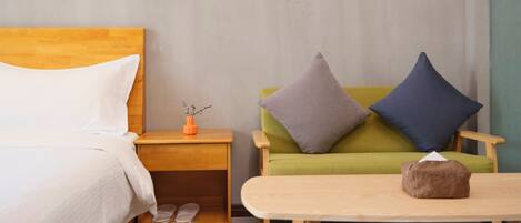 A neatly arranged hotel room with a bed, a wooden nightstand, a green sofa with two pillows, and a small wooden table with a tissue box.