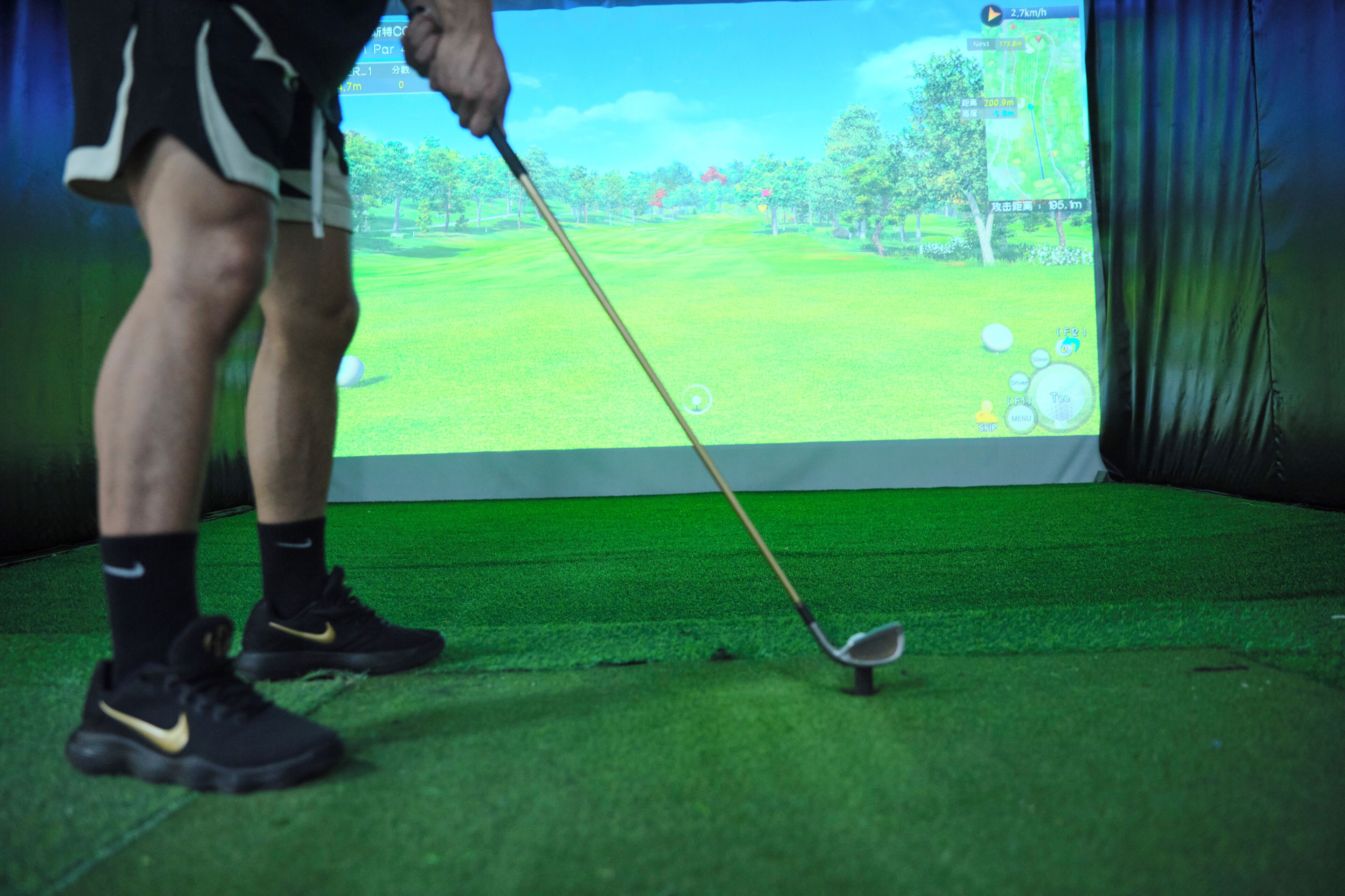 Una persona jugando golf sobre un césped verde, con un juego de golf virtual mostrado en una pantalla al fondo.