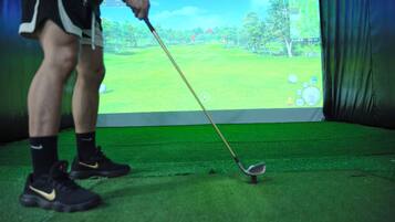 Una persona jugando golf sobre un césped verde, con un juego de golf virtual mostrado en una pantalla al fondo.