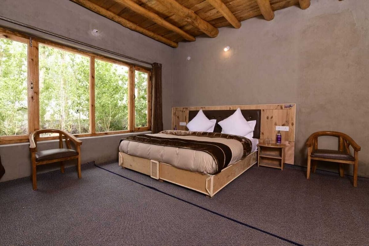 Un dormitorio con una cama grande, cabecera de madera, dos sillas, una mesita de noche y una ventana con vista a árboles.