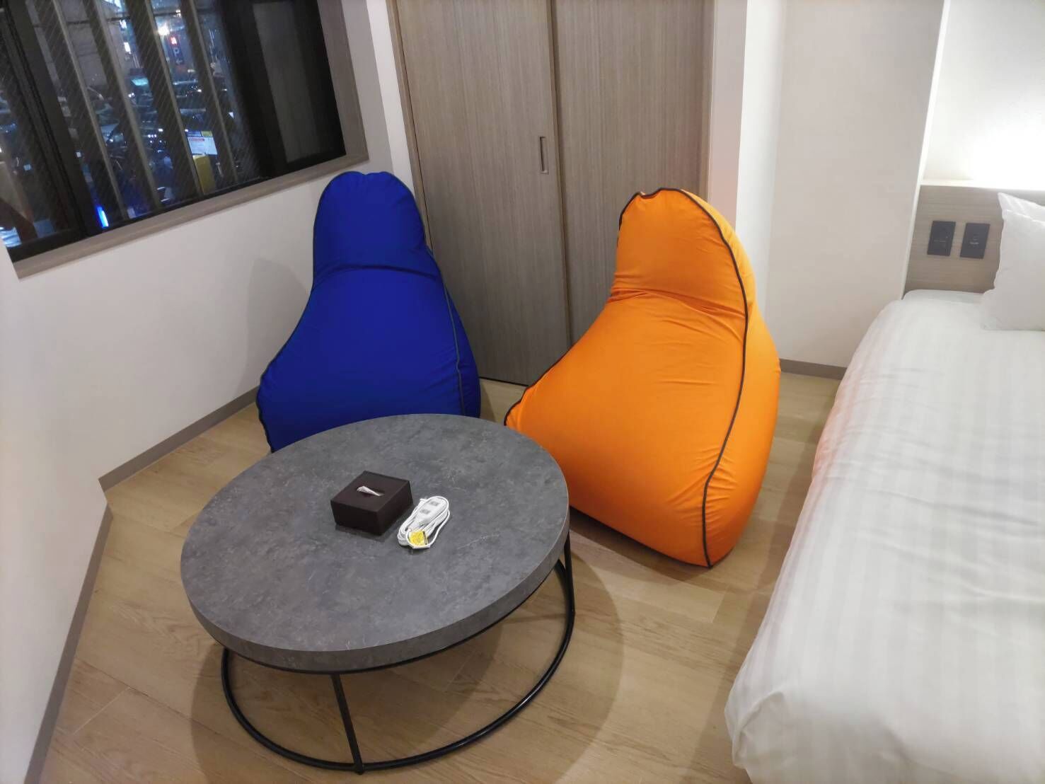 Una habitación pequeña y moderna con una mesa redonda, dos sillones de mimbre, una cama y una ventana con vistas a la ciudad.