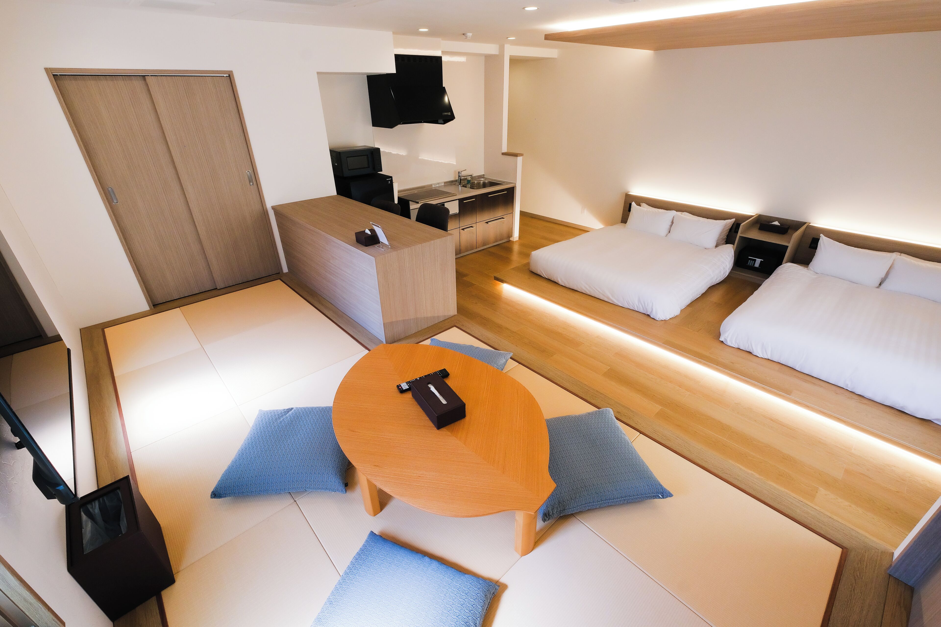 Una habitación de hotel moderna con suelo de madera, una pequeña mesa de comedor, dos camas, una kitchenette y un televisor montado en la pared.