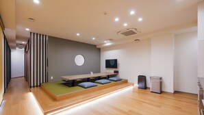 Una habitación moderna y bien iluminada con suelo de madera, una plataforma baja con cojines, un espejo redondo y un televisor en la pared.
