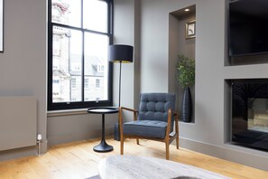 A modern living room with a large window, a standing lamp, a grey armchair, a small round table, and a fireplace. - Stylish 2-bed flat in New Town (Edinburgh)