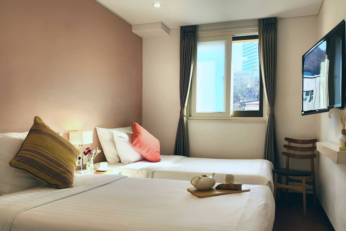 Une chambre d’hôtel avec deux lits, une fenêtre avec des rideaux, une chaise en bois et un téléviseur fixé au mur.