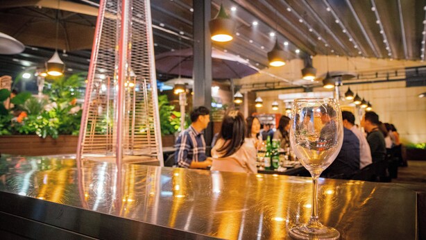 A restaurant interior with a bar area, hanging lights, and a large plant in the background.