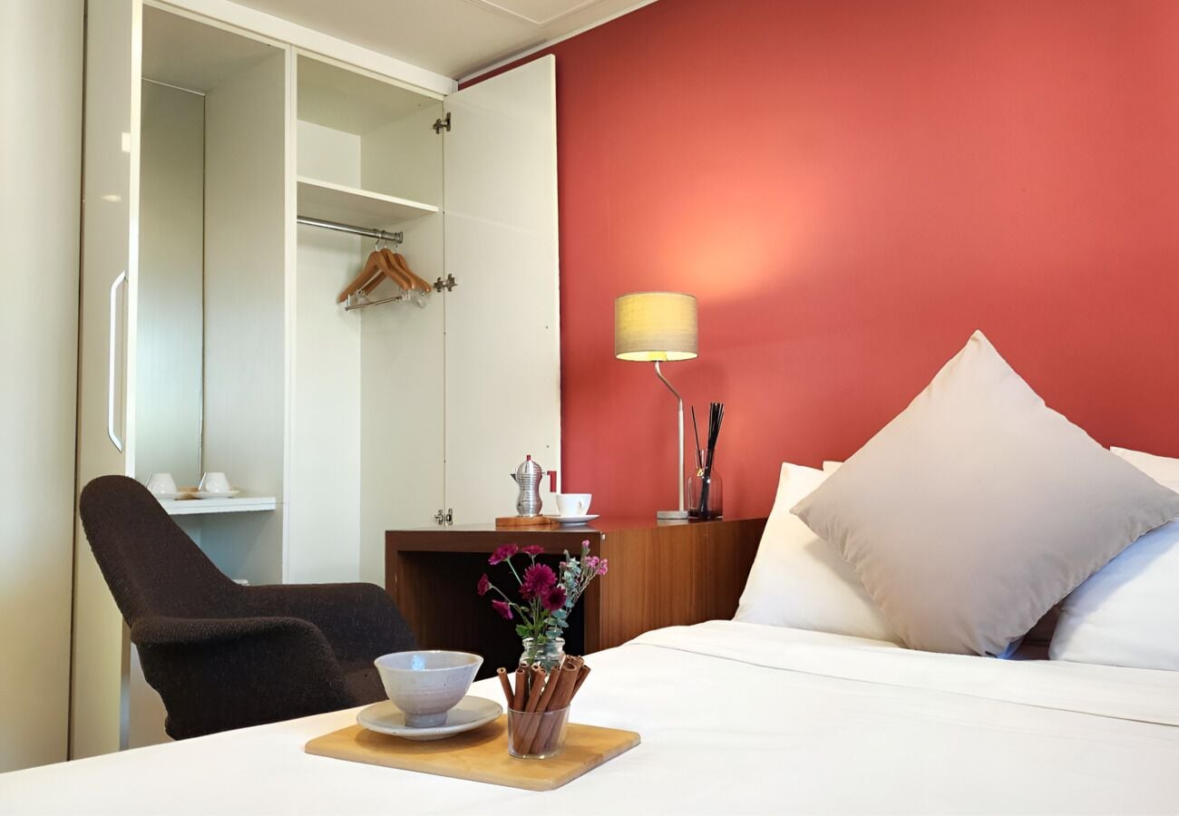 A hotel room with a bed, a chair, a bedside table with a lamp, a small table with a bowl and a vase of flowers, and a wardrobe.