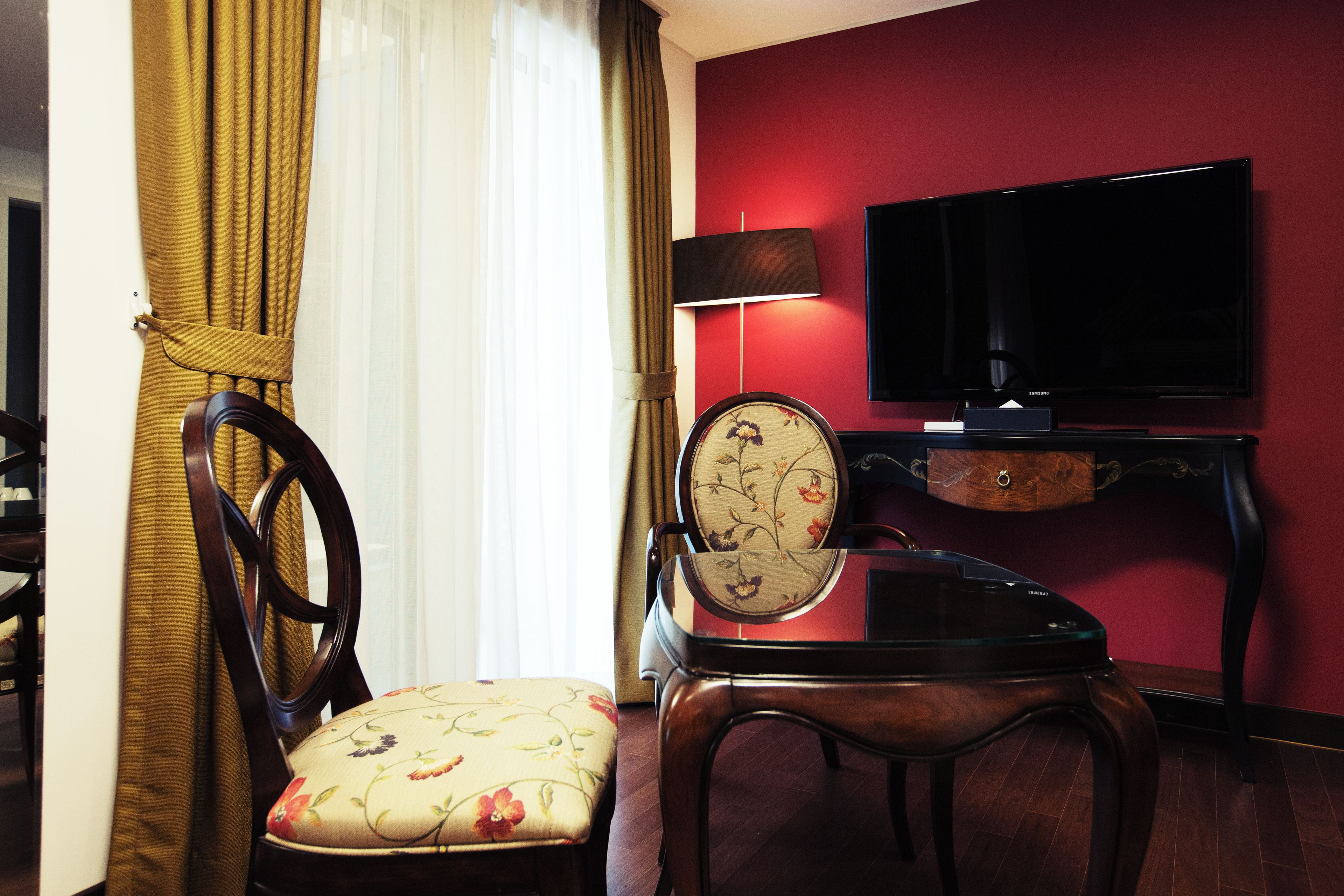 Une chambre d’hôtel avec une chaise en bois, une petite table, un téléviseur fixé au mur et une fenêtre avec des rideaux.