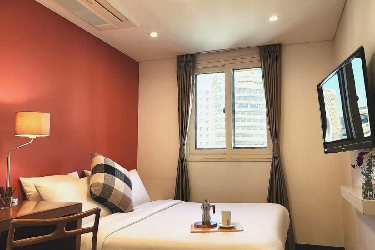 Une chambre d’hôtel avec un lit, un bureau doté d’une lampe, un téléviseur fixé au mur et une fenêtre avec des rideaux.