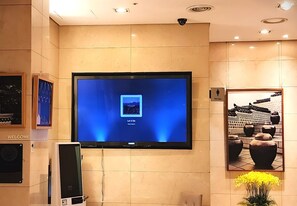 Zona de recepció d'hotel amb una pantalla gran que mostra un missatge de benvinguda, un cartell de 'Welcome' i una fotografia emmarcada d'arquitectura tradicional.