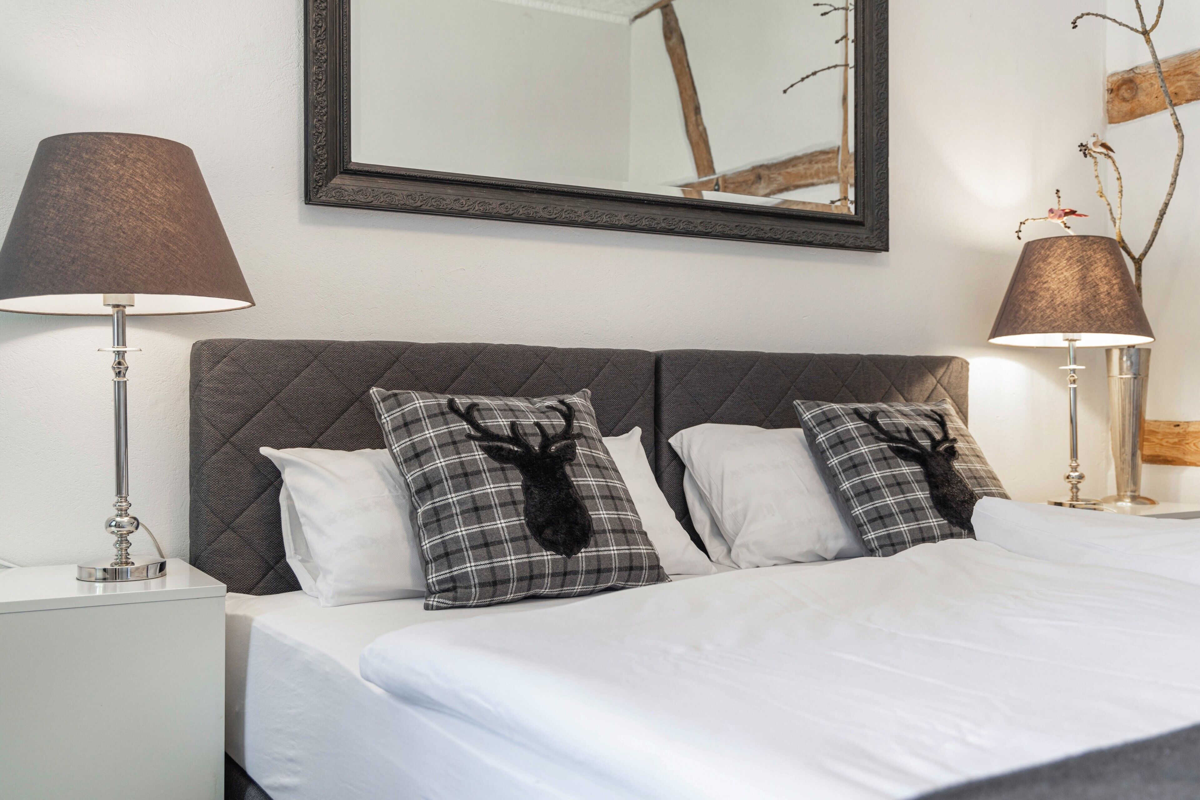 Een netjes opgemaakt bed met twee kussens, een bedlampje en een spiegel aan de muur.