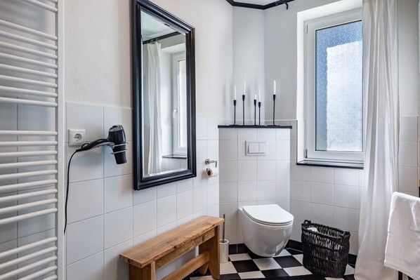 A bathroom with a white tiled floor, a wooden bench, a white toilet, a mirror, and a window with a view.