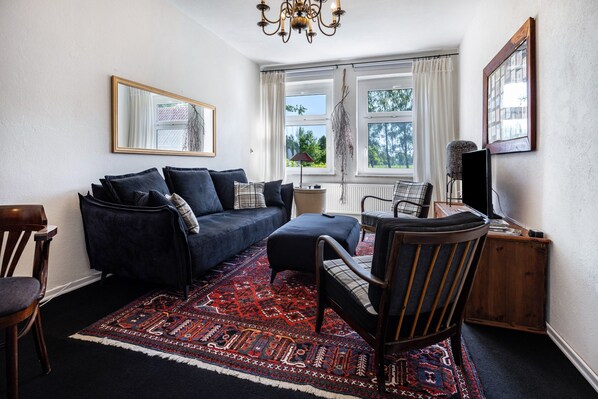 A living room with a dark blue sofa, a wooden chair, a red patterned rug, and a large window with curtains. - Holiday Apartment 'Landhaus Heiddorf' with 2 Comfortable Bedrooms, Terrace with BBQ & Sun Loungers (Neu Kaliß)