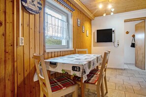 Een kamer met houten lambrisering, een eettafel, stoelen, een klok, een raam met gordijnen, een wandtelevisie en een deur.