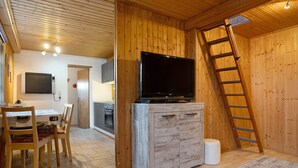 A wooden interior with a TV on a cabinet, a dining area with chairs, and a kitchen in the background.