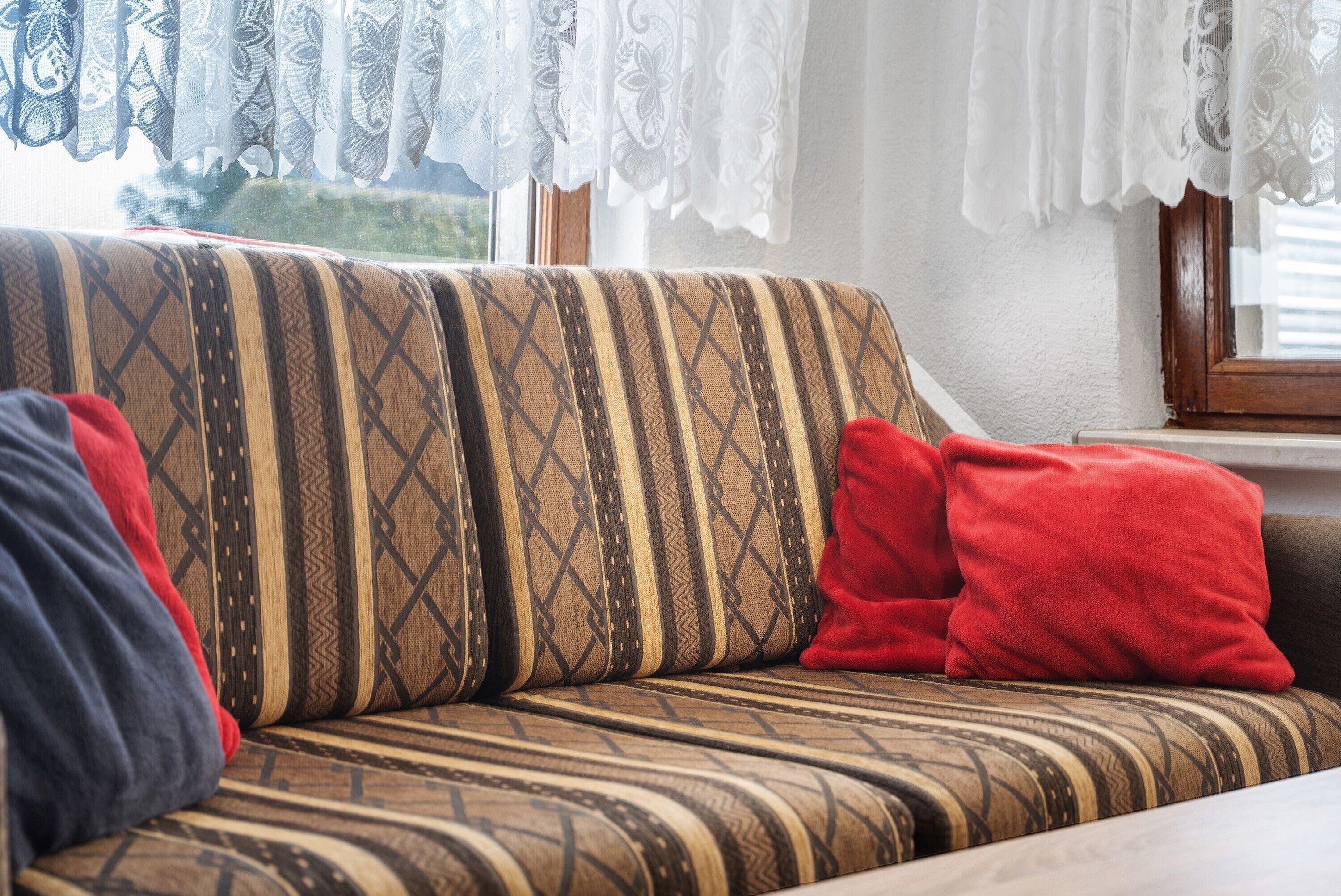 Un canapé recouvert d’un tissu à motifs, agrémenté de coussins rouges et bleus, placé près d’une fenêtre ornée de rideaux en dentelle.