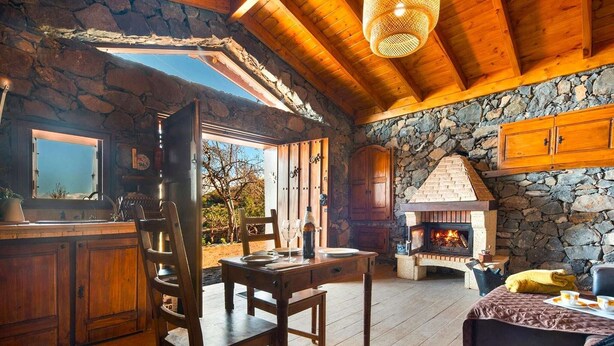 A rustic cabin interior with a stone fireplace, wooden furniture, and a large window overlooking a garden.