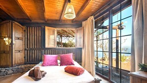 A wooden cabin bedroom with a large bed, a window with a view of the outdoors, and a hanging light fixture.
