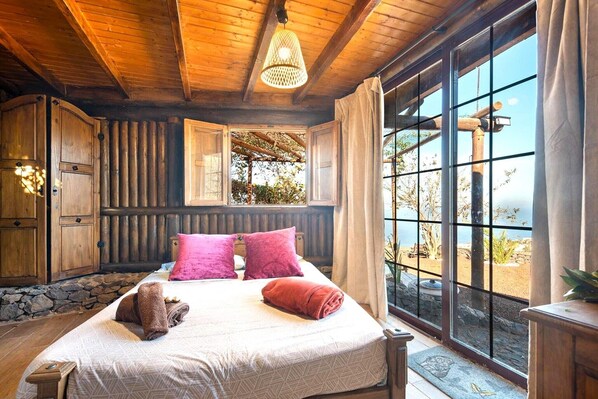 Un dormitorio de cabaña de madera con una cama grande, una ventana con vistas al exterior y una lámpara colgante.