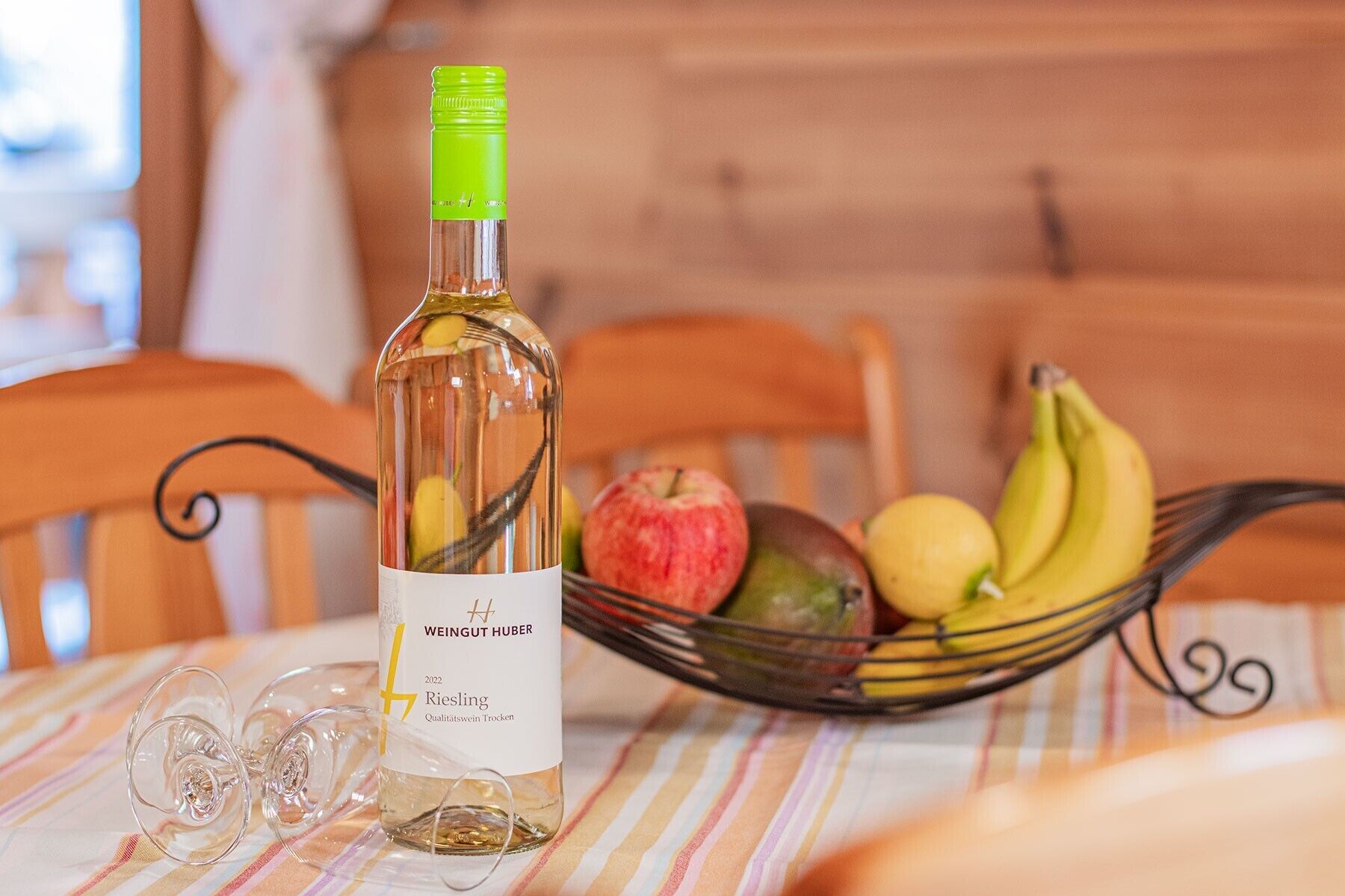 A bottle of Riesling wine, a glass, and a fruit bowl with apples, bananas, and lemons on a table.