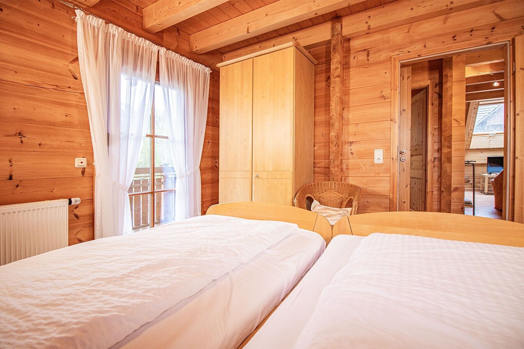 Ein Zimmer in einer Holzhütte mit zwei Betten, einem Korbstuhl und einem großen Fenster mit Vorhängen.