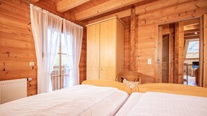 A wooden cabin room with two beds, a wicker chair, and a large window with curtains.