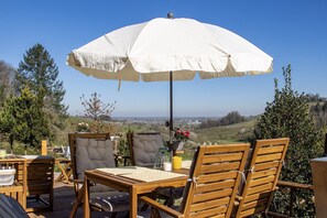 A wooden deck with a table and chairs, a white umbrella, and a view of a landscape.