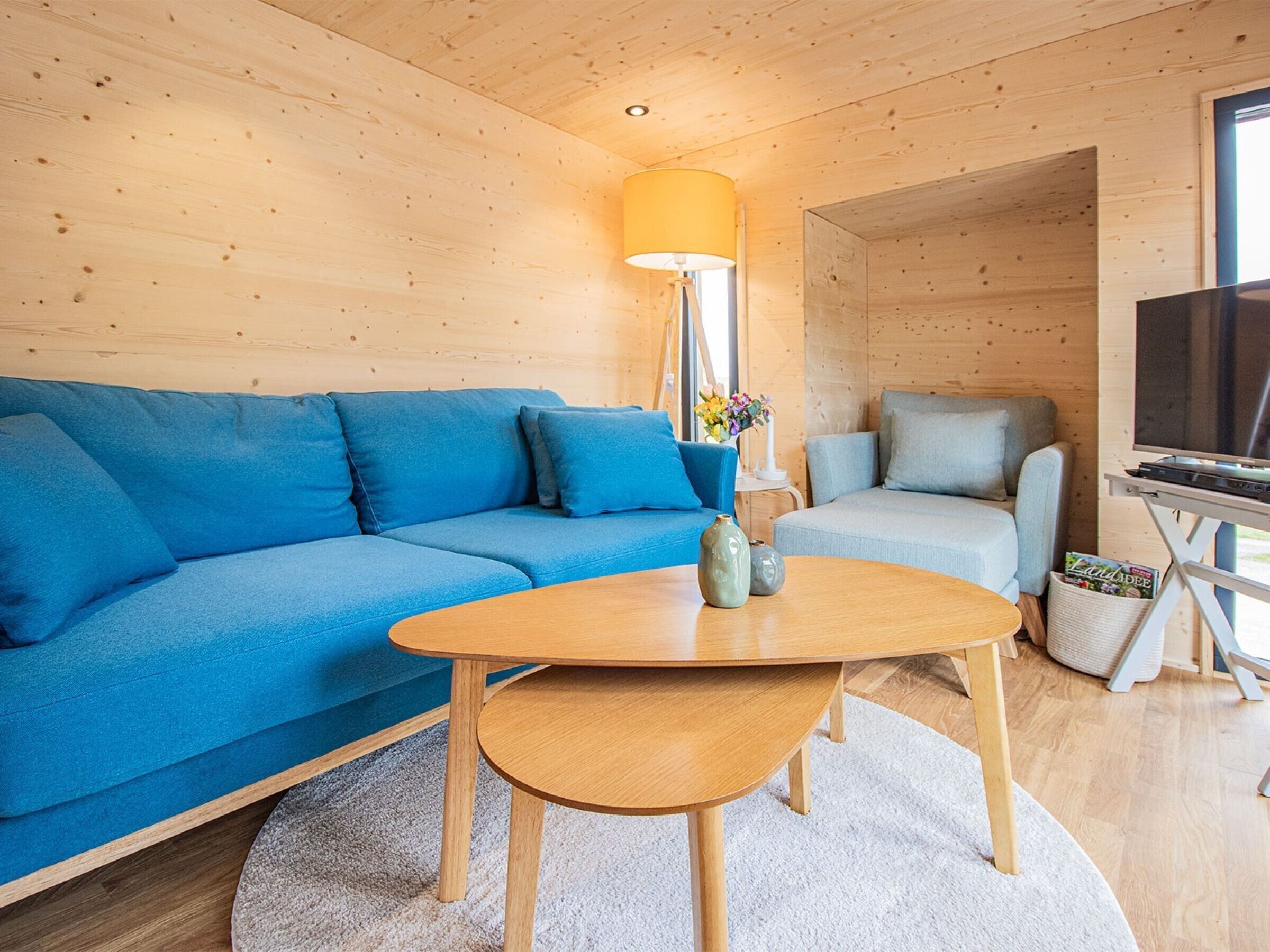 A wooden interior with a blue sofa, a grey armchair, a wooden coffee table, a lamp, and a television.