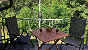 Et træbord med stole på en udendørs terrasse, omgivet af et metalgelænder, med en frodig grøn baggrund.