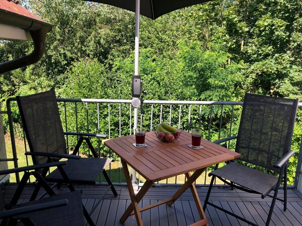 A wooden table with chairs on an outdoor deck, surrounded by a metal railing, with a lush green background.