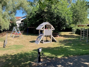 Une aire de jeux avec un toboggan, des balançoires et une petite maison en bois.