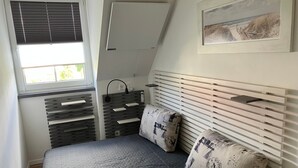 Et soveværelse med en seng, et vindue med skodder, en fastmonteret varmeblæser, et natbord og et billede i ramme på væggen.