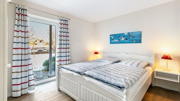 A bedroom with a bed, curtains, a painting, two bedside lamps, and a view through a window. - Apartment 'Stine' with Shared Terrace, Shared Garden and Wi-Fi (Breklum)