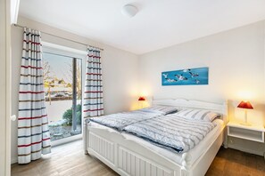Ein Schlafzimmer mit einem Bett, Vorhängen, einem Gemälde, zwei Nachttischlampen und Blick durch ein Fenster.