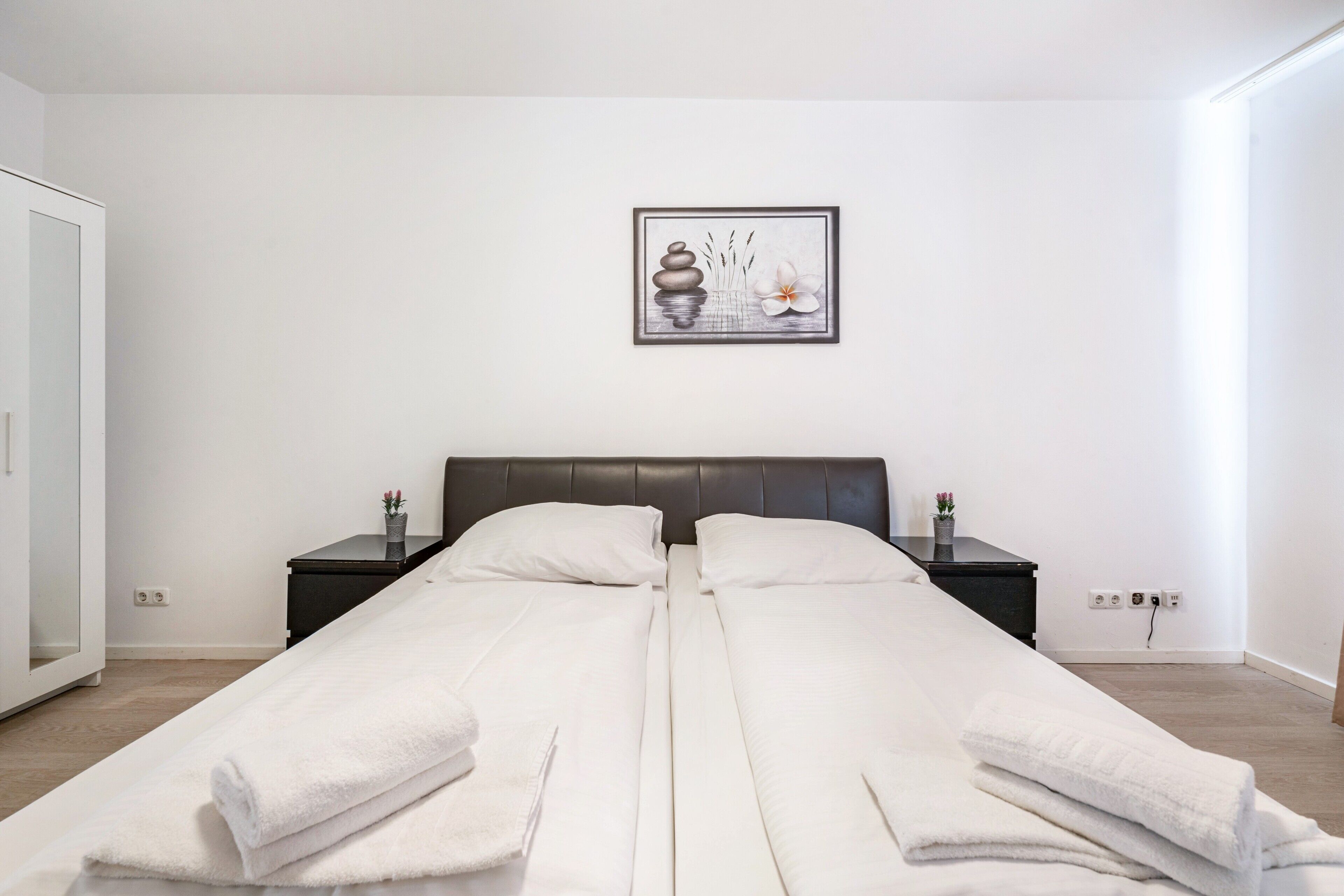 A double bed with white linens, a dark headboard, bedside tables with lamps, a framed artwork on the wall, and a white door.