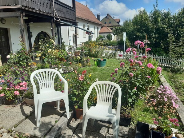 A garden with two white plastic chairs, a white building with a red roof, and a white picket fence.