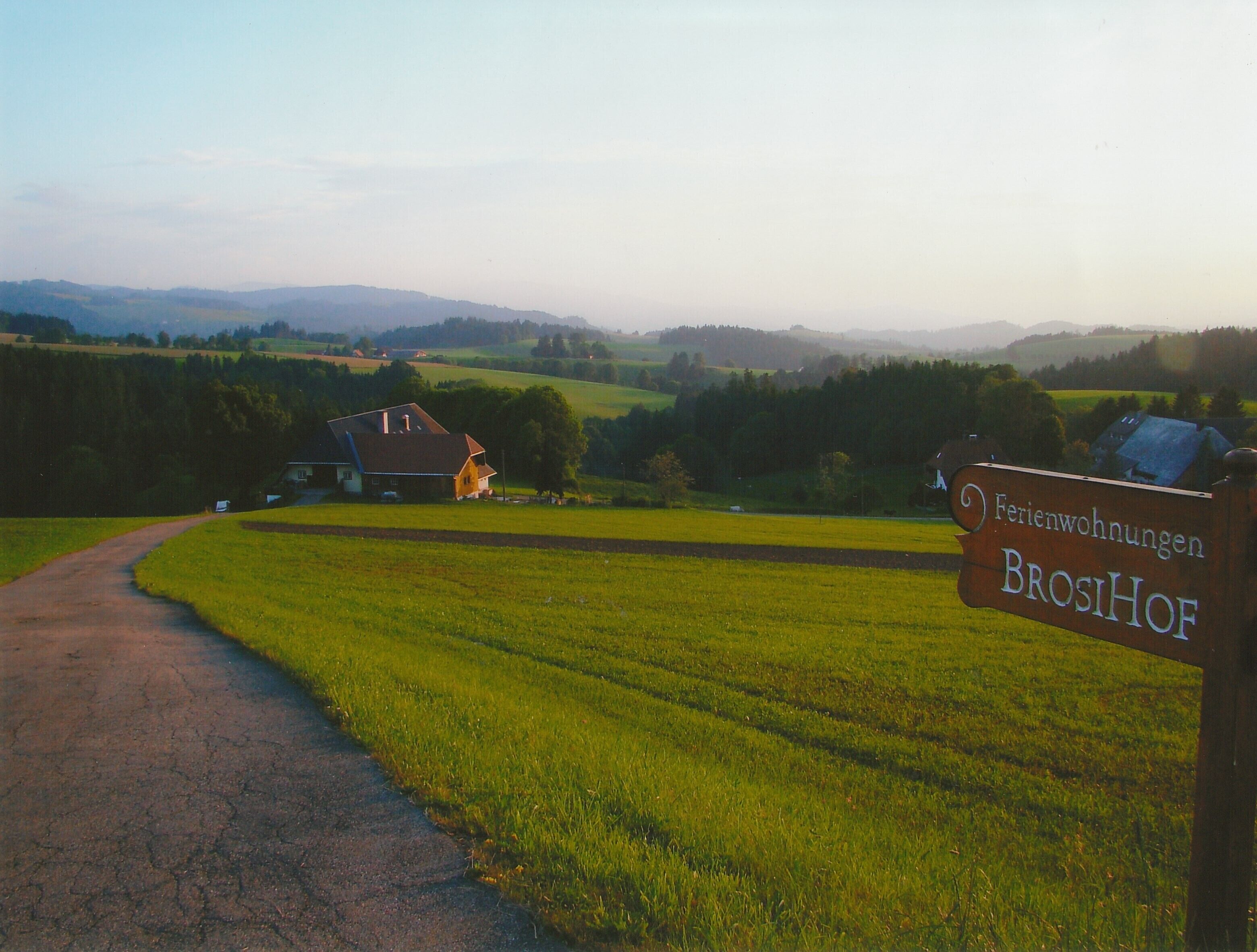 Ländliche Landschaft mit einer Feldstraße, einem Haus und einem Schild mit der Aufschrift 'Ferienwohnungen BROSIOF'.