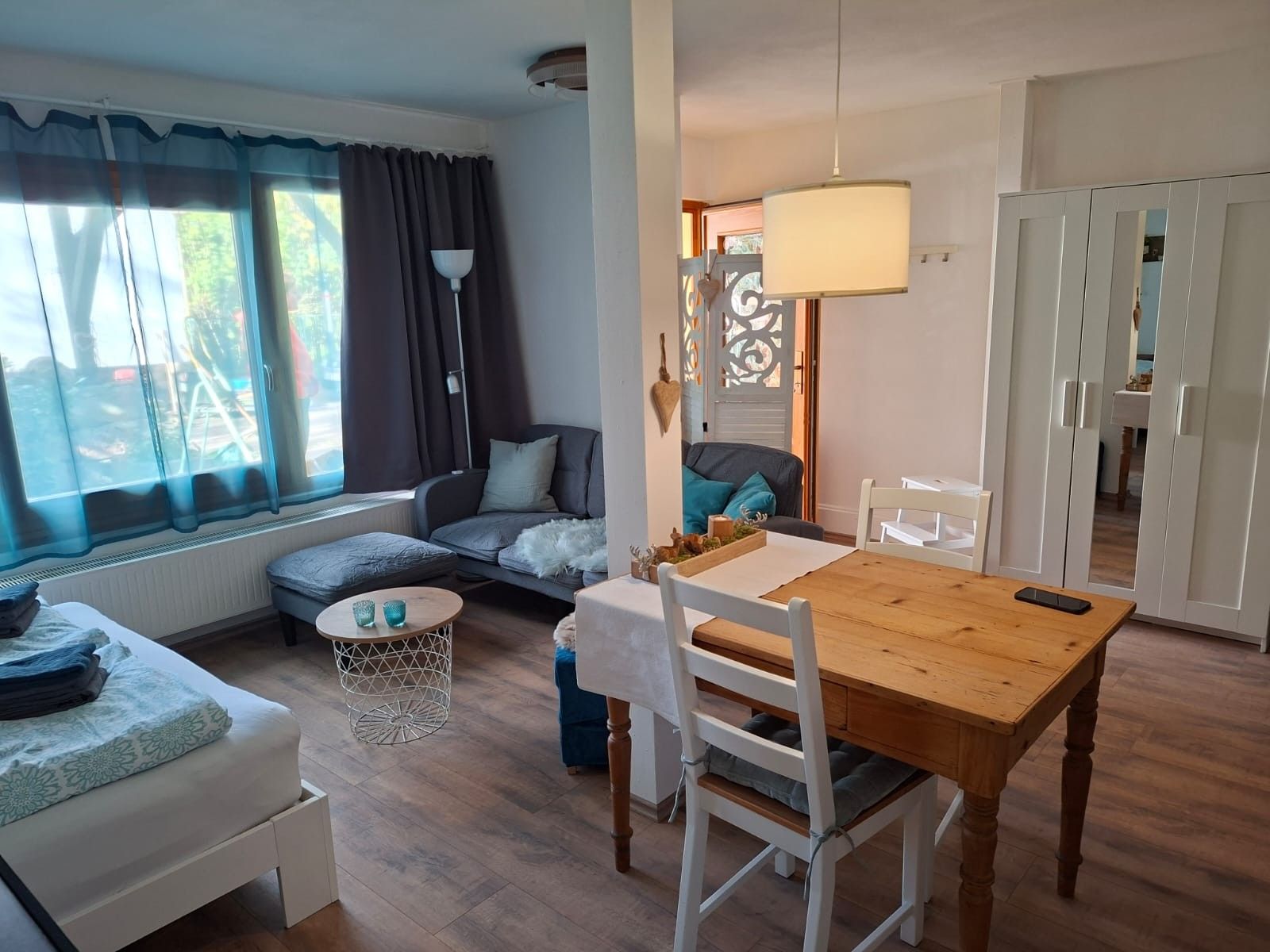A cozy living room with a sofa, a dining table with chairs, a small round table with a lamp, and a window with curtains.
