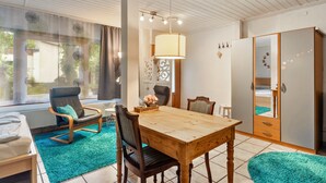 A room with a wooden dining table, two chairs, a sofa, a window with curtains, a lamp, and a wardrobe.