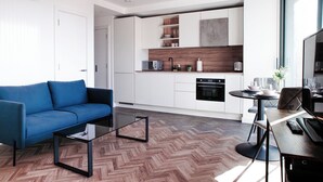 A modern living room with a blue sofa, a glass coffee table, and a dining area with a table and chairs. The kitchen features white cabinets, a wooden countertop, and a built-in oven. - Horizon: A Modern Luxury Studio (London)