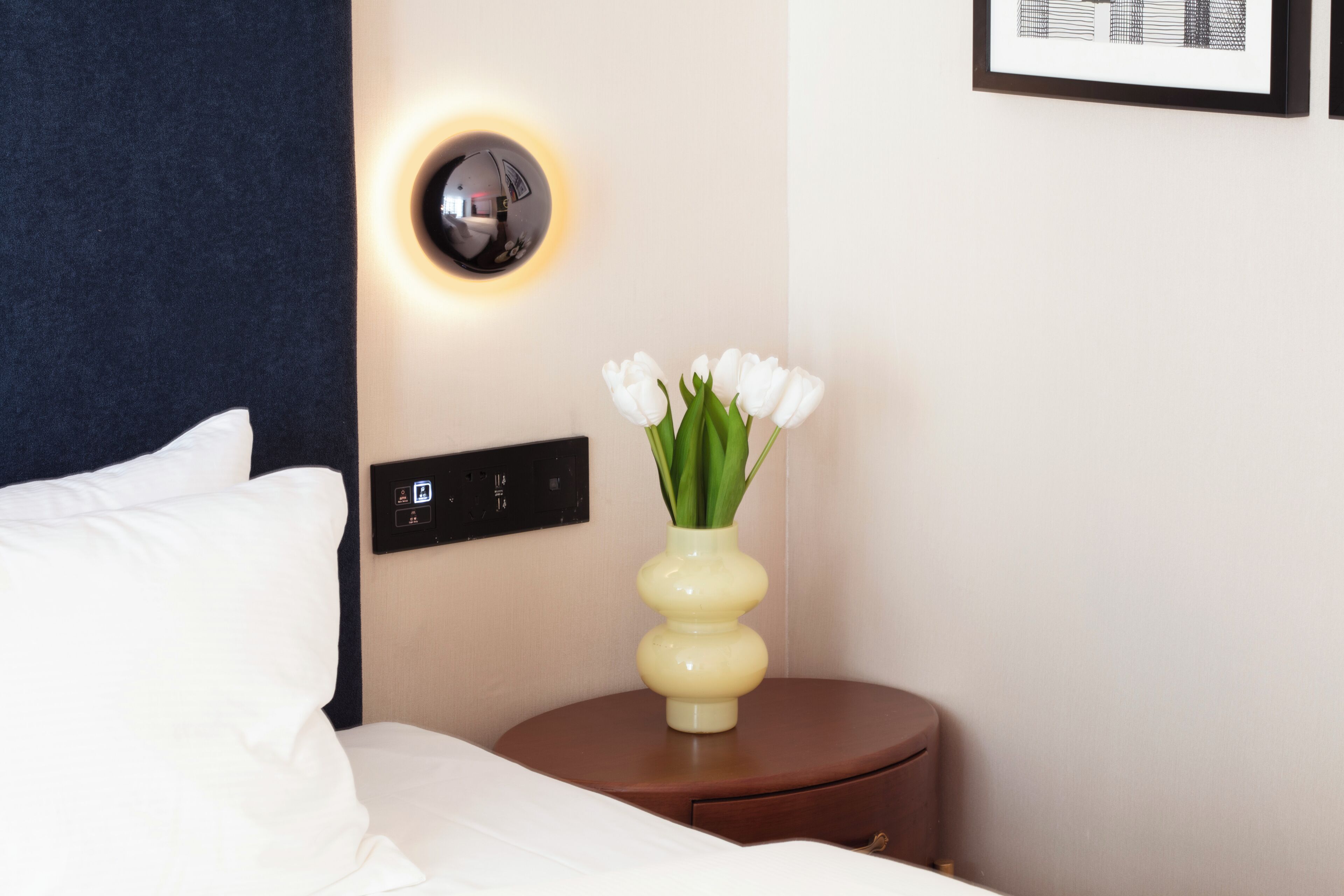 A neatly made bed with white linens, a bedside table with a vase of white flowers, a wall-mounted light fixture, and a framed picture on the wall.