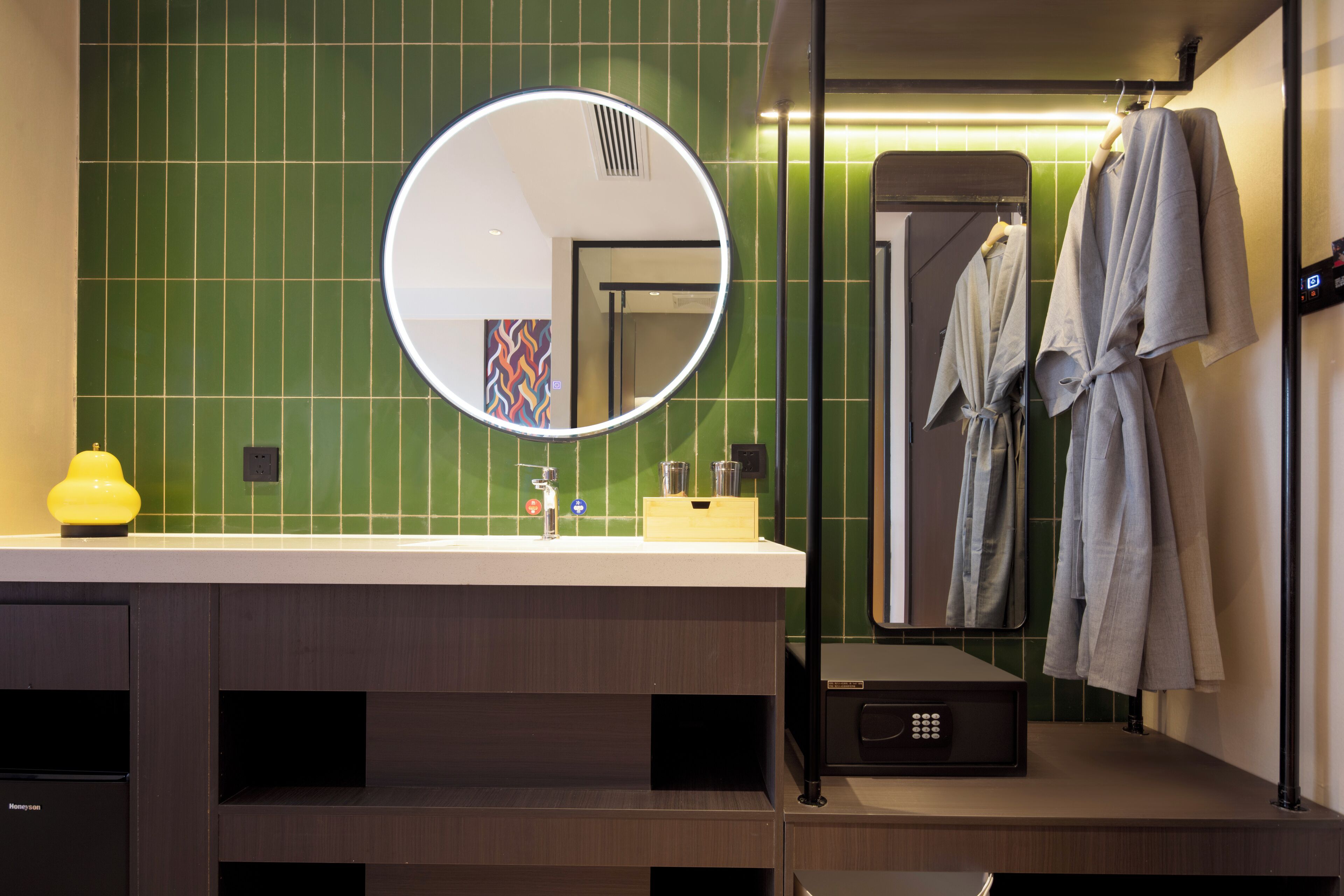 A modern bathroom with a green tiled wall, a round mirror, a sink with a yellow soap dispenser, and a robe hanging on a rack.