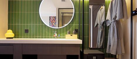 A modern bathroom with a green tiled wall, a round mirror, a sink with a yellow soap dispenser, and a robe hanging on a rack.