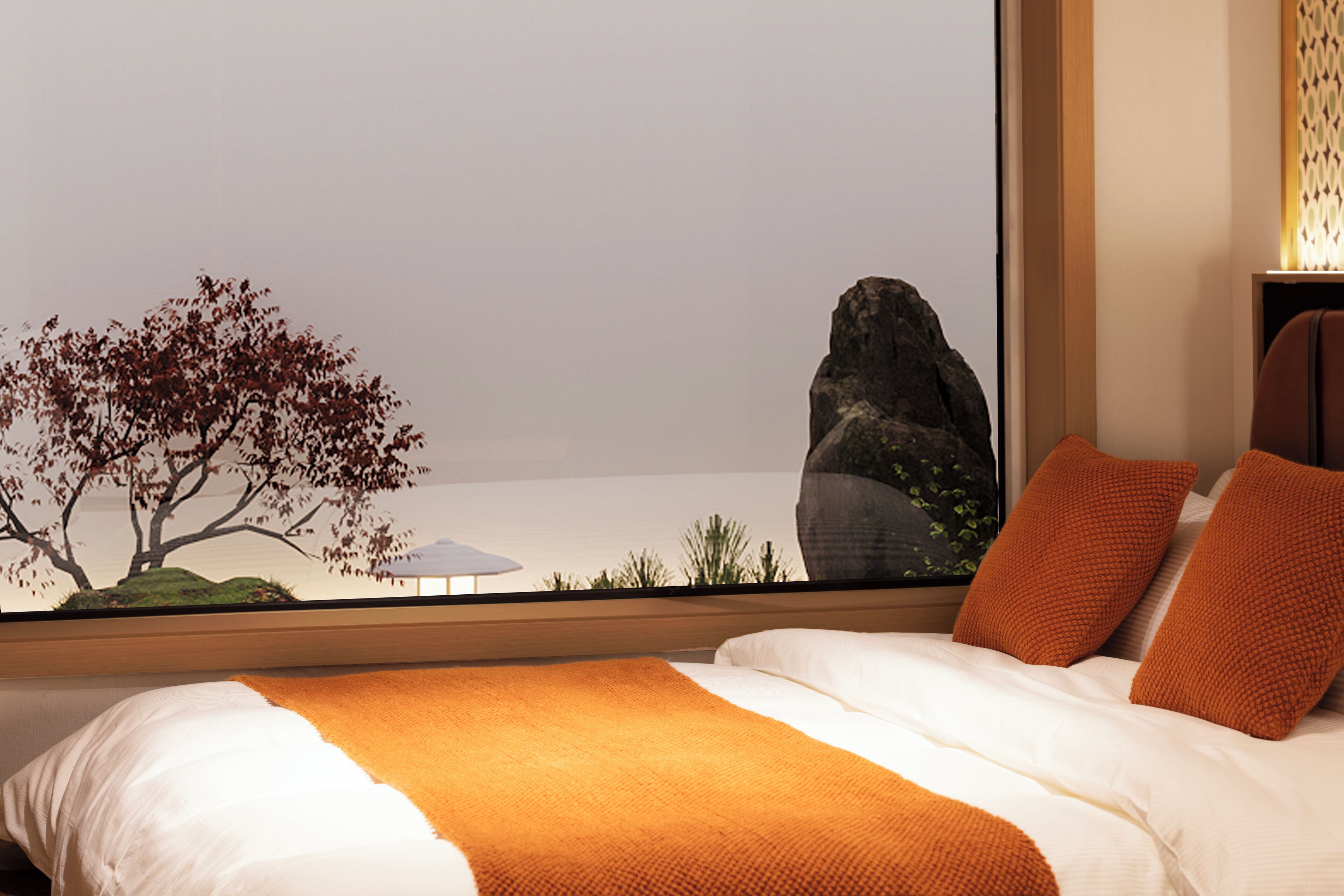 A bedroom with a large bed, orange bedding, a view of a tree and rock through a window, and a nightstand with a lamp.