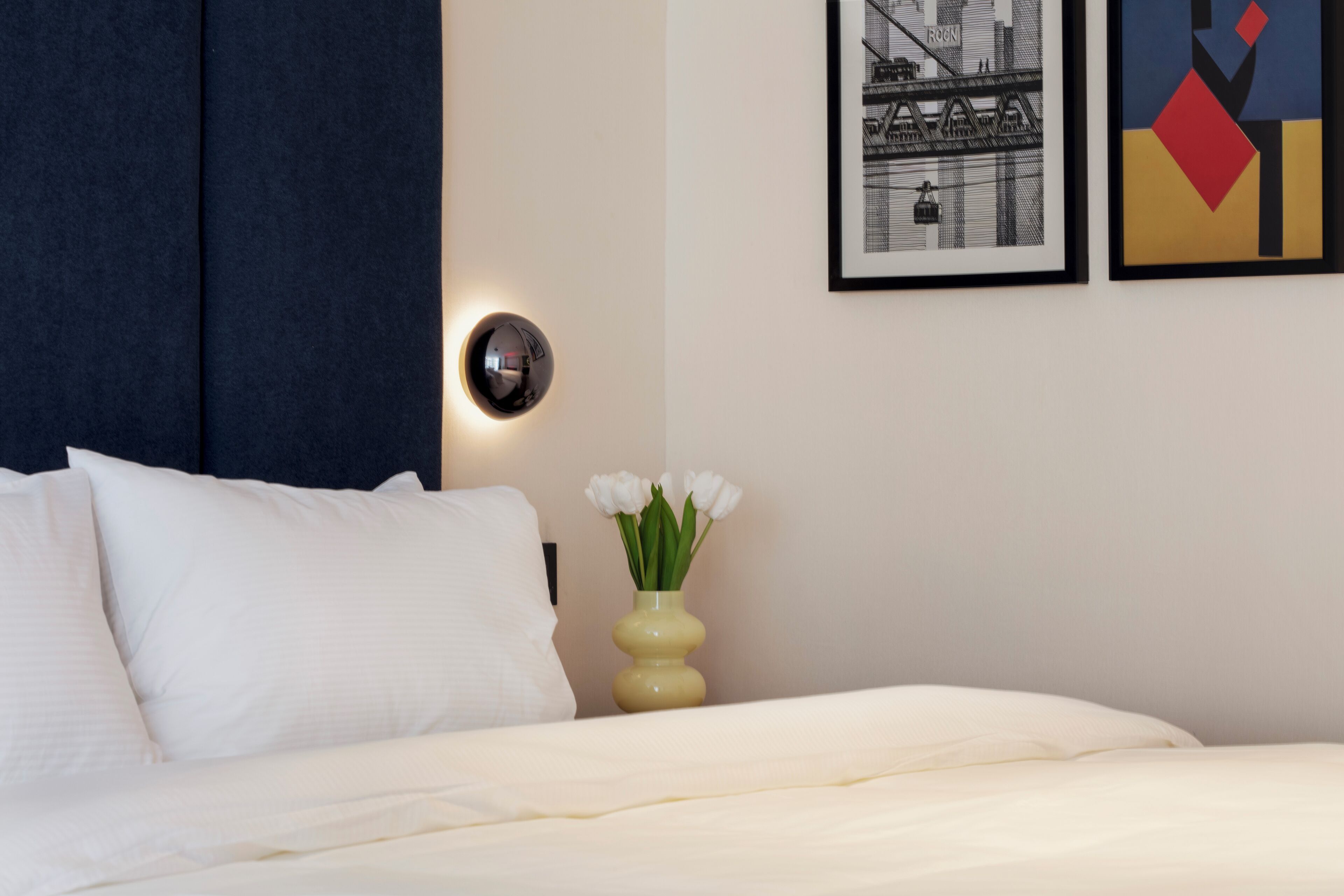 A neatly made bed with white linens, a vase with white flowers, and two framed pictures on the wall.
