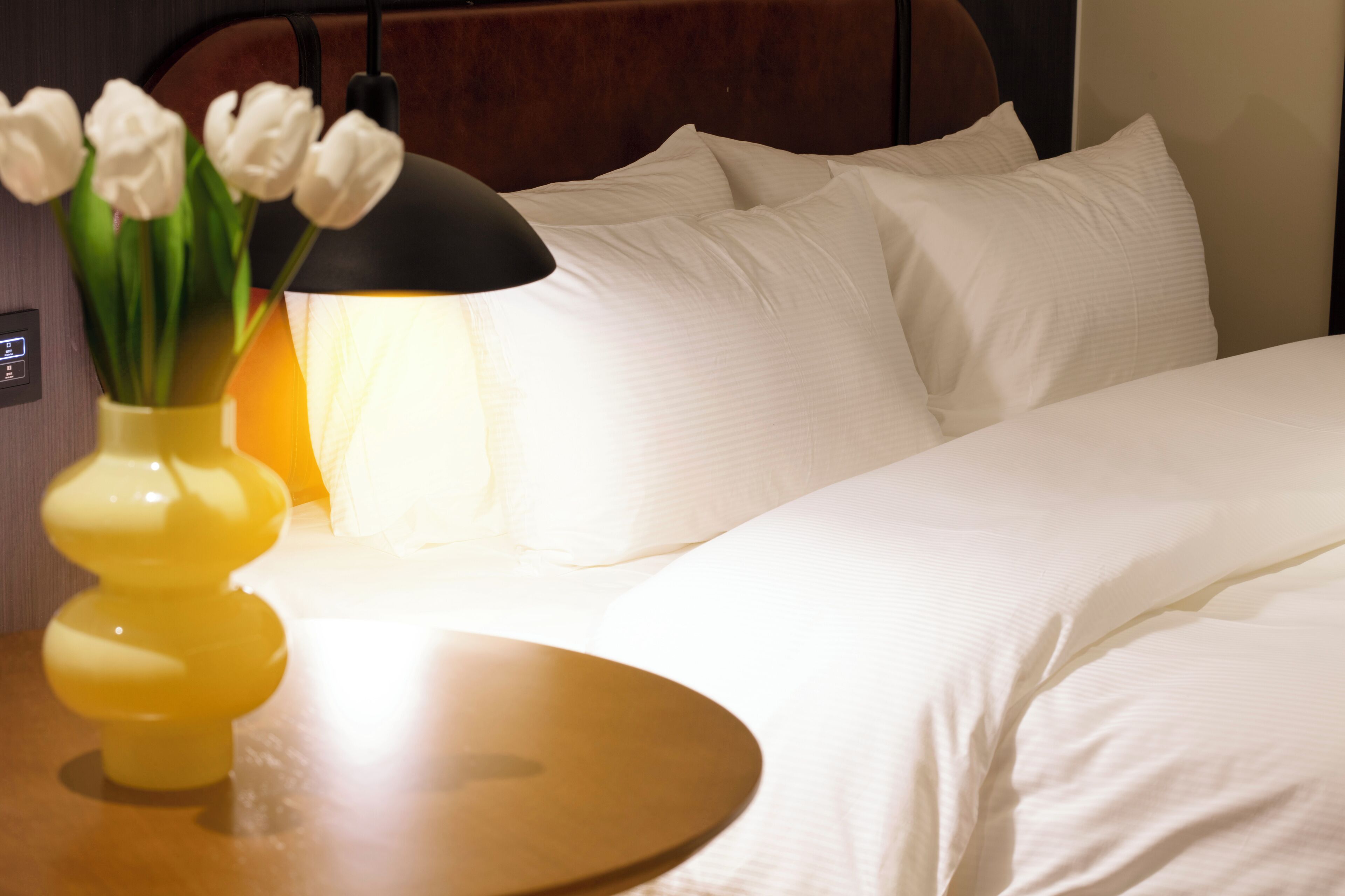 Een keurig opgemaakt bed met witte lakens, een nachtkastje met een gele vaas en witte tulpen, en een lamp met een zwarte lampenkap.