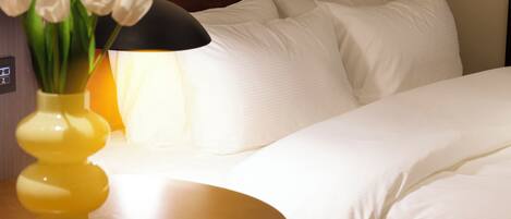 Een keurig opgemaakt bed met witte lakens, een nachtkastje met een gele vaas en witte tulpen, en een lamp met een zwarte lampenkap.