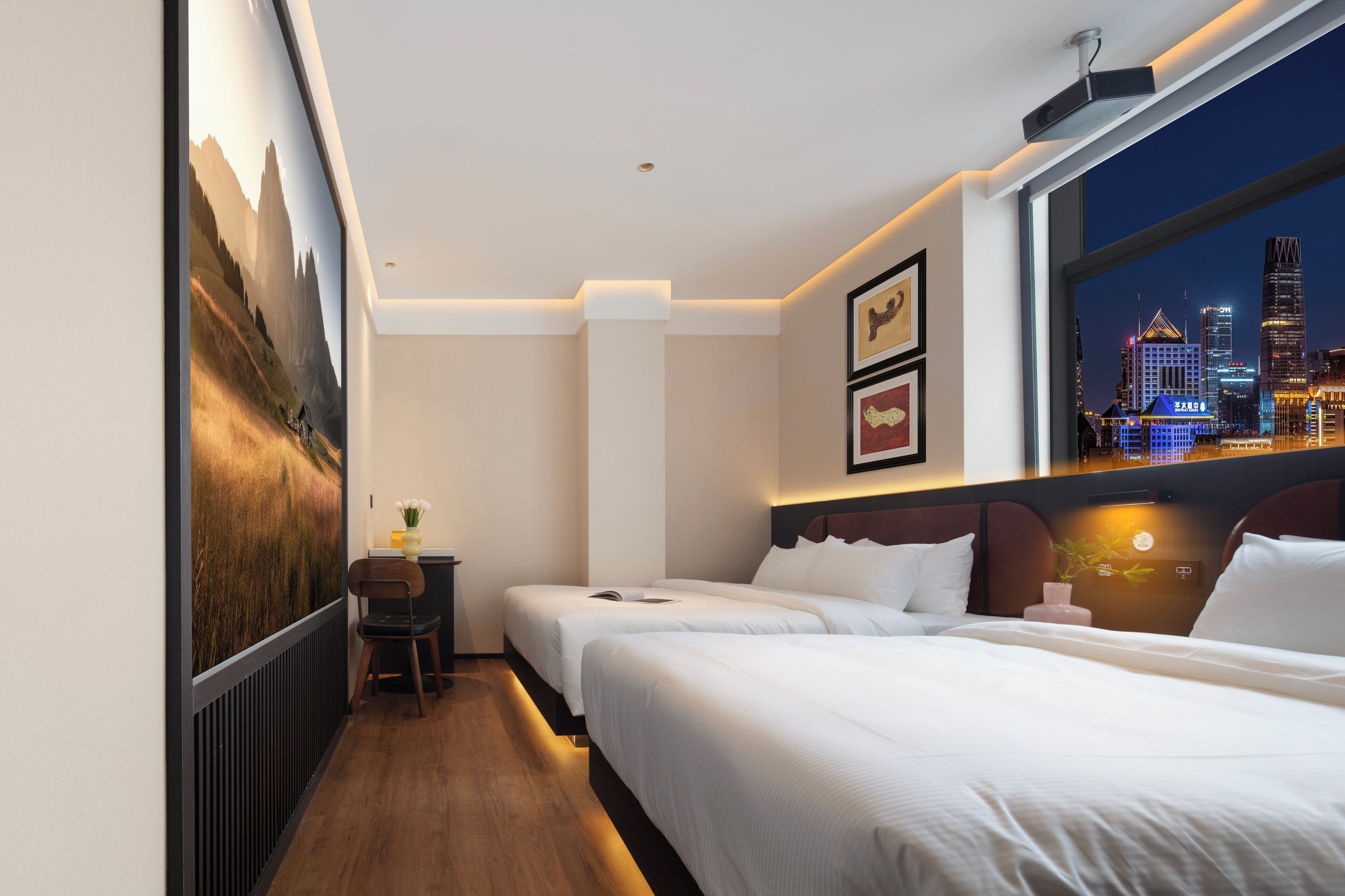 Een hotelkamer met twee bedden, uitzicht over de stad, een lijst met foto aan de muur en een klein tafeltje met een stoel.